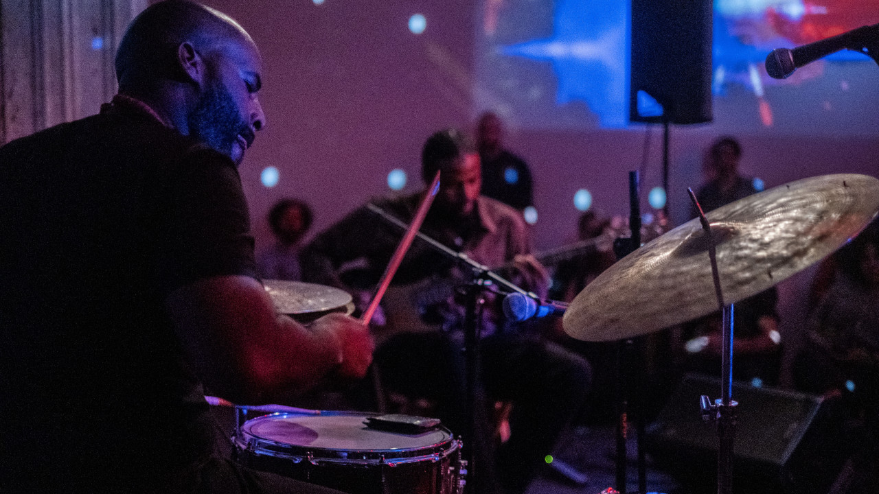 Two figures musicians perform on stage. On the foreground, a figure is playing the drums. On the background sits a guitar player.  Blue and red lights are projected on the back wall.