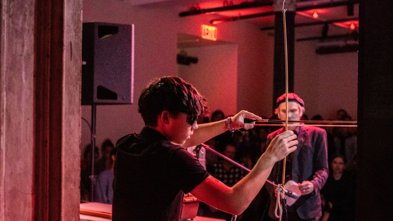 Close-up color image of a figure with a violin bow on their left hand. A thin, long piece of wood hangs from the ceiling, and the figure is using the bow to play it like a intrument string. The bow and the wood make a cross, which falls exactly in front of the face of a second figure. Also a performer, they are holding a white object in their hands. In the background, audience members. The space is illuminated by red light.