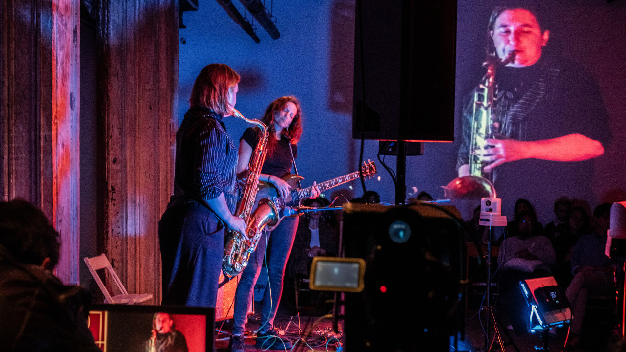 A color image of two figures performing on a stage. Various electronic equipment, such as speakers, cables, an monitors surround the performers. One of the figures is playing the saxophone. The other one is playing the guitar and has their eyes closed. An image of the figure playing the saxophone is projected onto the back wall. In the background, a row of audience members sit looking towards the performers.