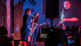 A color image of two figures performing on a stage. Various electronic equipment, such as speakers, cables, an monitors surround the performers. One of the figures is playing the saxophone. The other one is playing the guitar and has their eyes closed. An image of the figure playing the saxophone is projected onto the back wall. In the background, a row of audience members sit looking towards the performers.
