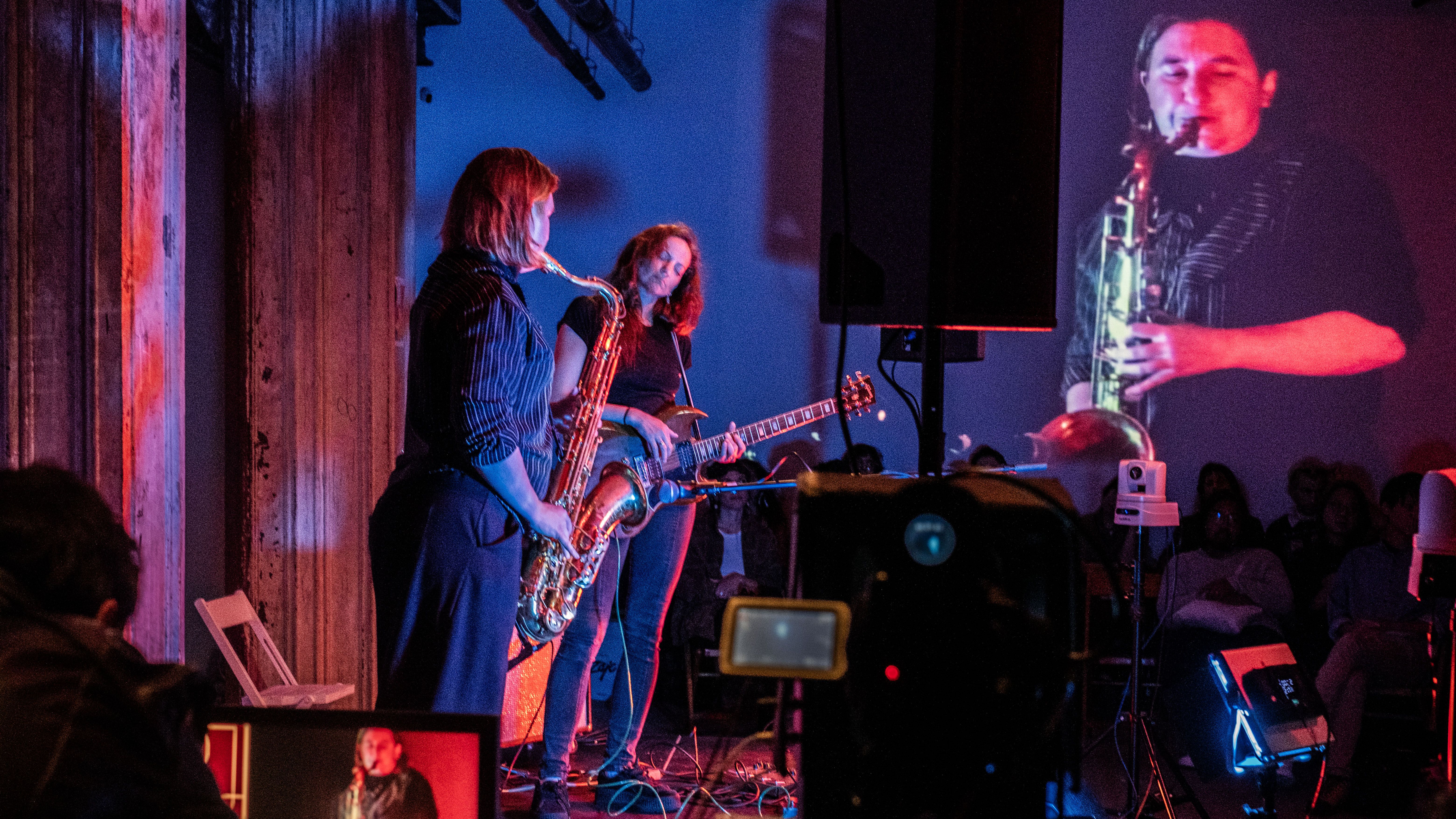 A color image of two figures performing on a stage. Various electronic equipment, such as speakers, cables, an monitors surround the performers. One of the figures is playing the saxophone. The other one is playing the guitar and has their eyes closed. An image of the figure playing the saxophone is projected onto the back wall. In the background, a row of audience members sit looking towards the performers.