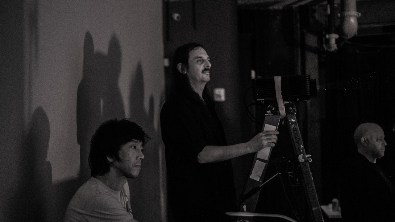 A black and white image of three figures. All three of them attentively look to the right of the image, towards the same direction. The figure in the center, holds a ladder, which has a projector taped to the top, with their right hand.