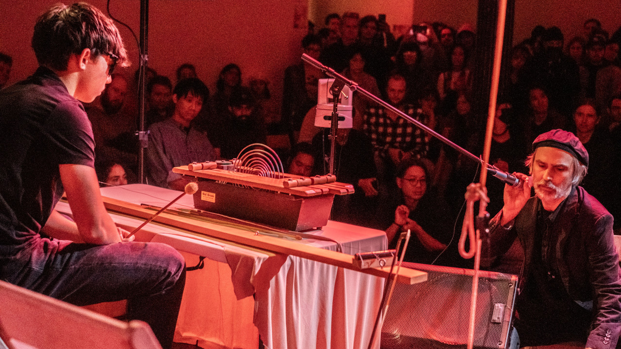 Color image of two figures facing each other, performing. The figure on the left holds a ramrod on his left hand, and is sitting on a chair. In front of the figure, there is a table with unusual instruments made of wood. The second figure is on the right. They are holding a microphone. In the background, audience members face the performers. The room is illuminated by red light.