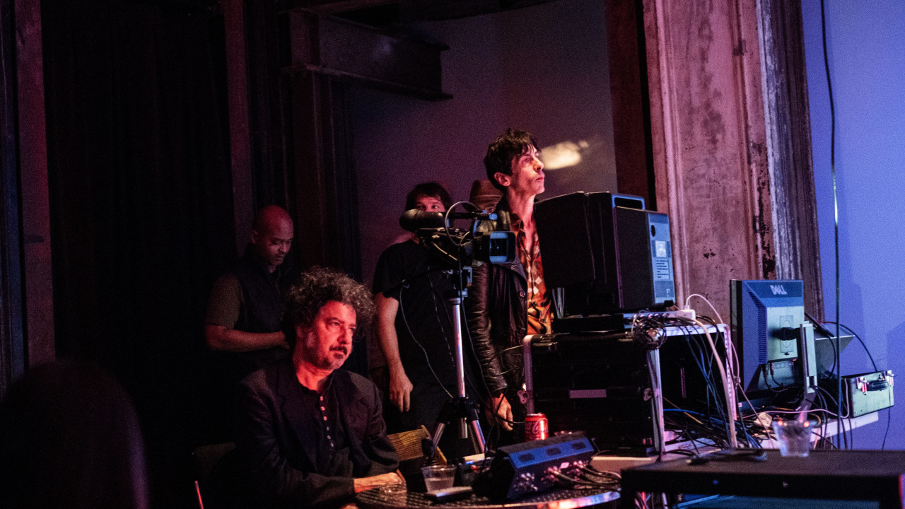 A color image of two figures before a table filled with electronic equipment such as cables, cameras, and monitors. The figure on the left is sitting down, and the figure on the right is standing up. They are both looking at something outside of the frame. Their faces are illuminated by red light.