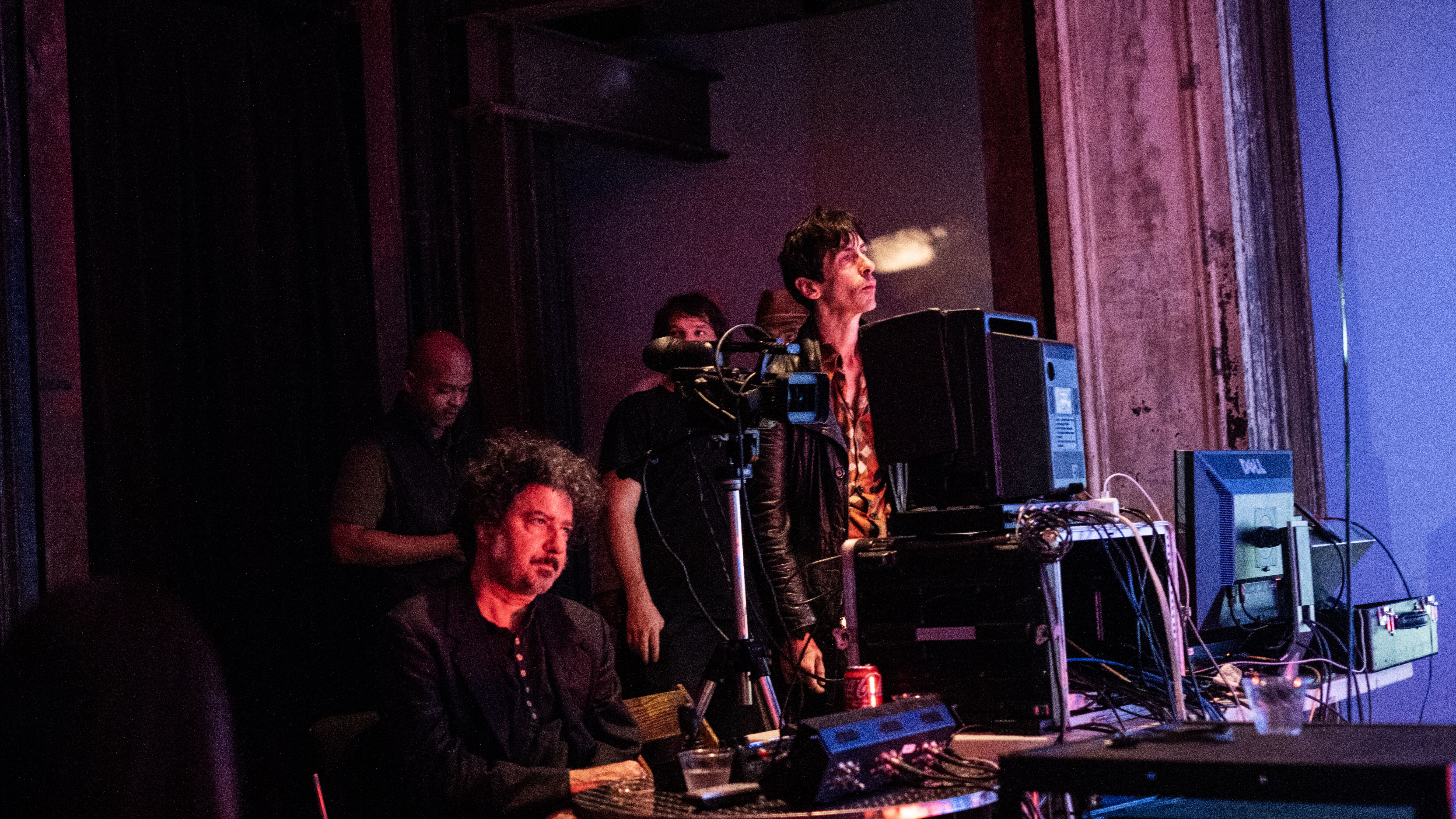 A color image of two figures before a table filled with electronic equipment such as cables, cameras, and monitors. The figure on the left is sitting down, and the figure on the right is standing up. They are both looking at something outside of the frame. Their faces are illuminated by red light.