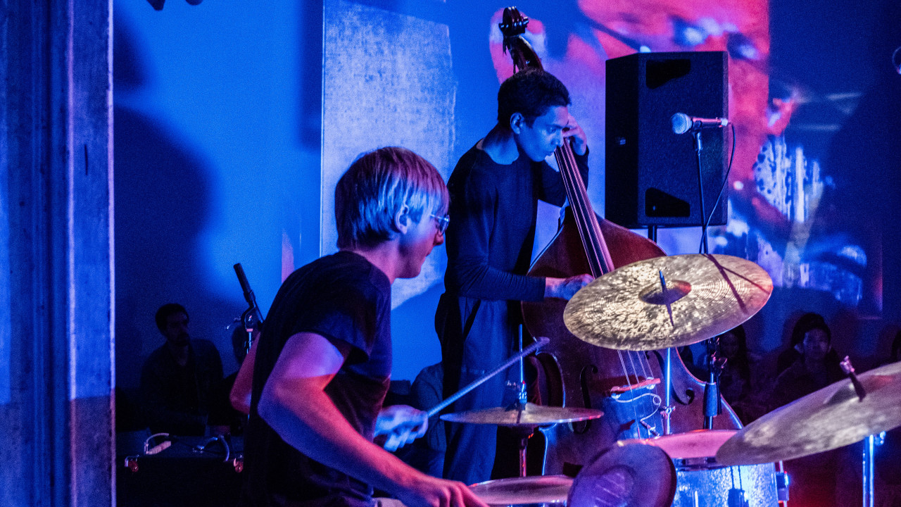 A close-up color image of two figures performing. One plays the drums, and the second figure plays the double bass. To their right, a large black speaker, and a microphone. Images are projected onto the back wall. The room is illuminated with red and blue light.