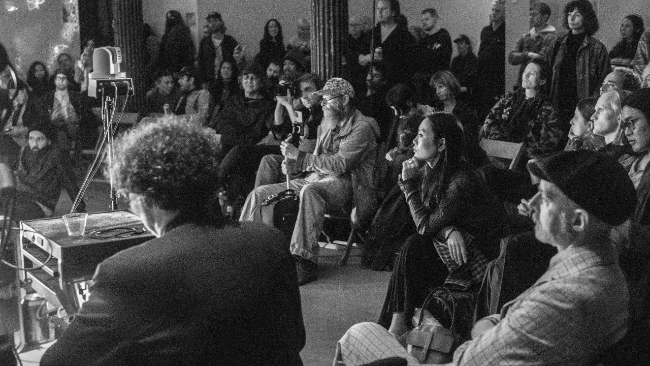 Black and white image of an audience watching a performance. Some are sitting on chairs, and others stand.