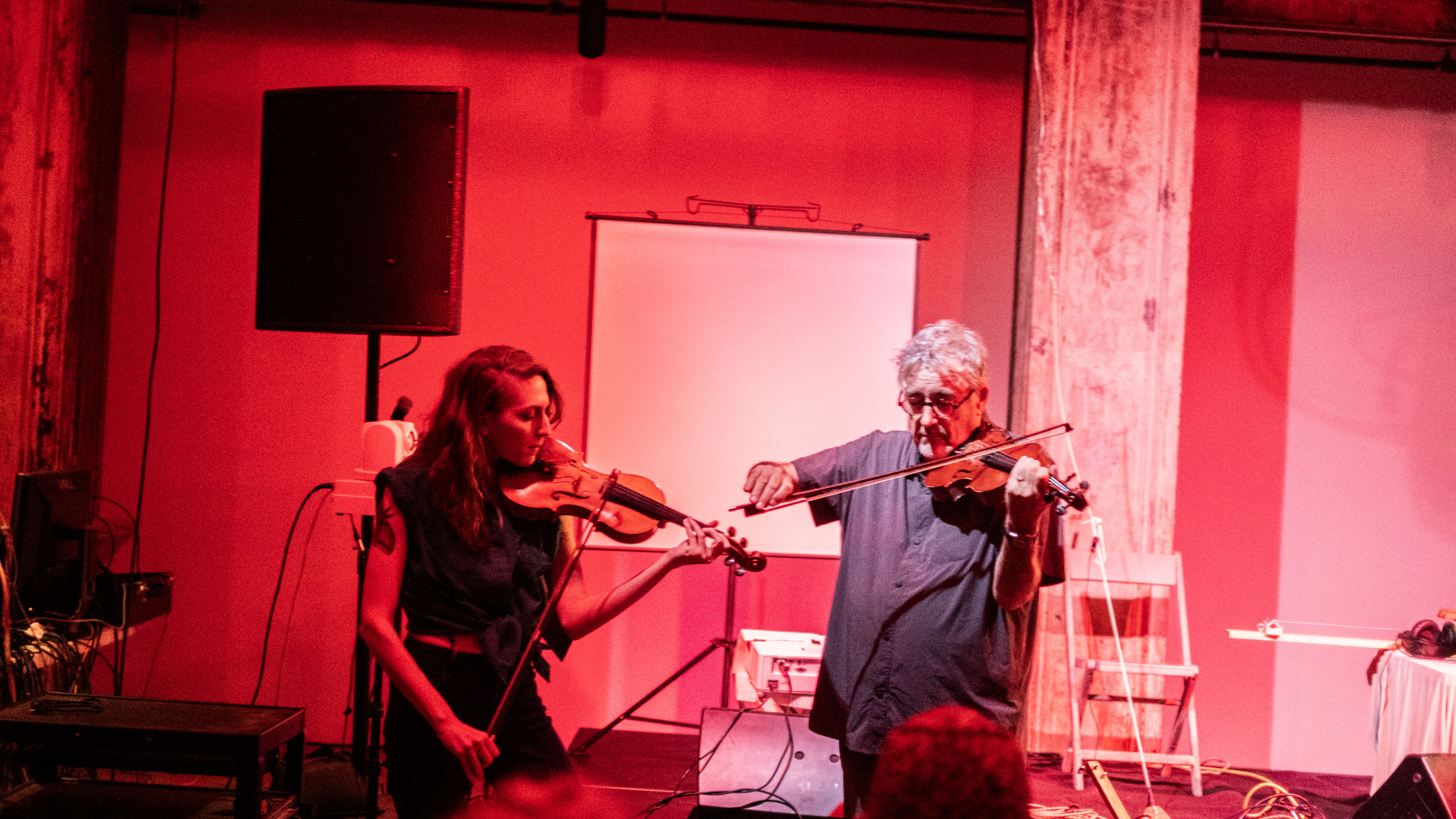Color image of a performance space illuminated by red light. In the center, two figures standing next to each other are playing the violin. Behind them is a white projector screen and a speaker.