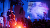 Color image of a performance space illuminnated with blue and red light. On the left, a figure on a stage plays the double bass, there is a large black speaker to their right.  Abstract images are projected onto the wall. Below the projections, audience members sit on chairs.