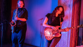 A color image of two figures performing. The figure on the left plays the saxophone. There is a standing microphone in front of them. The figure on the right, has their eyes closed, and is playing the guitar. Blue and red light is projected onto them.