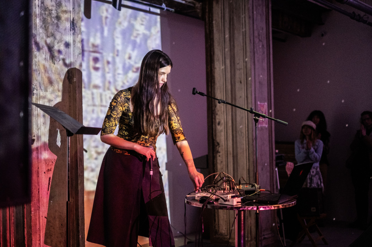 Color image of performer using their left hand to control a soundboard resting on a circular table while their left hand grasps a microphone. Next the performer is an empty music stand. The performer