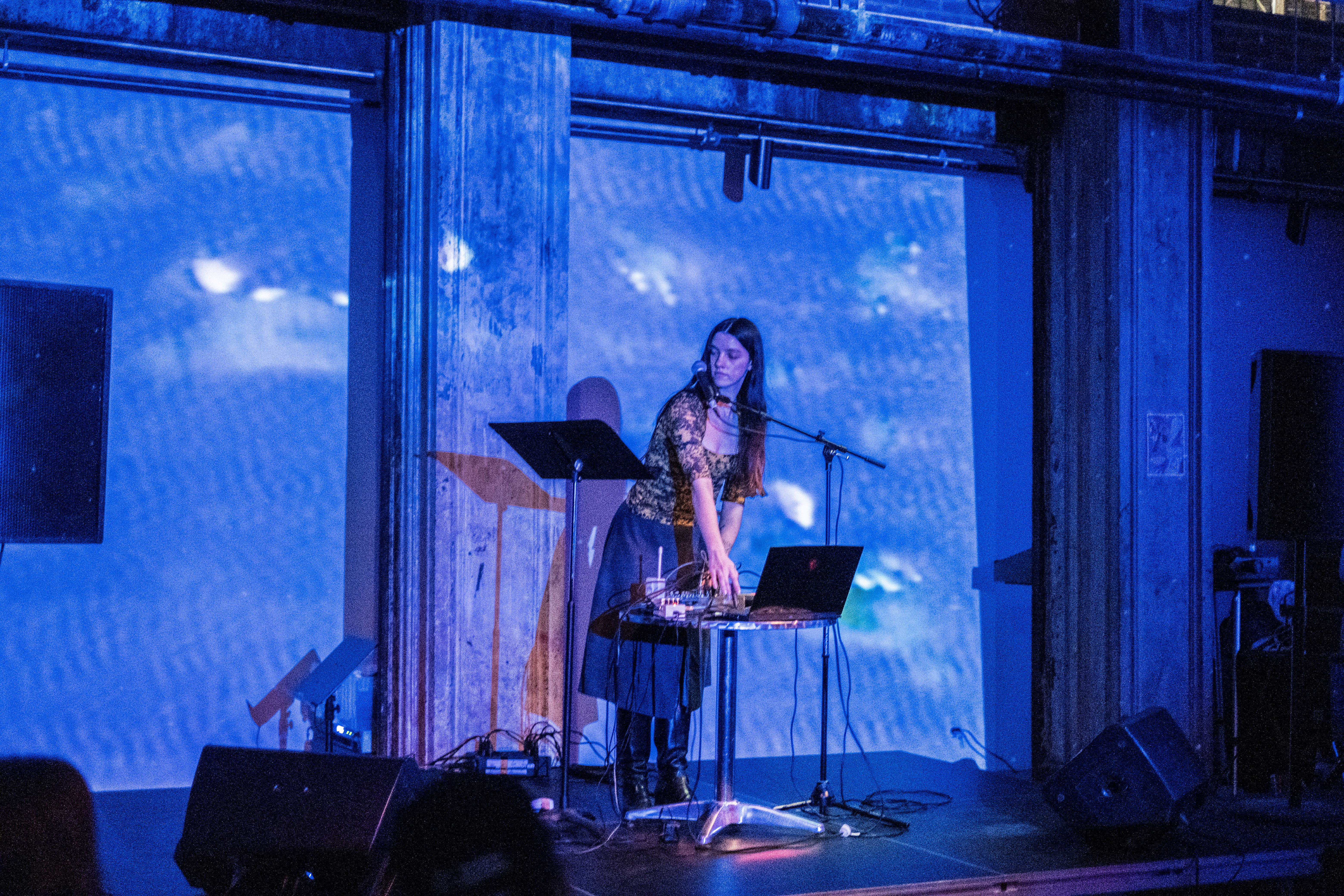 Color image of a performer standing on stage in front of a circular table. The performer bends over slightly to play music from a laptop and soundboard. Their face leans into a microphone while looking stage right at a music stand placed next to the circular table. A blue light emitted from a projector covers the whole stage and casts shadows of the performer and their setup onto the back wall. 