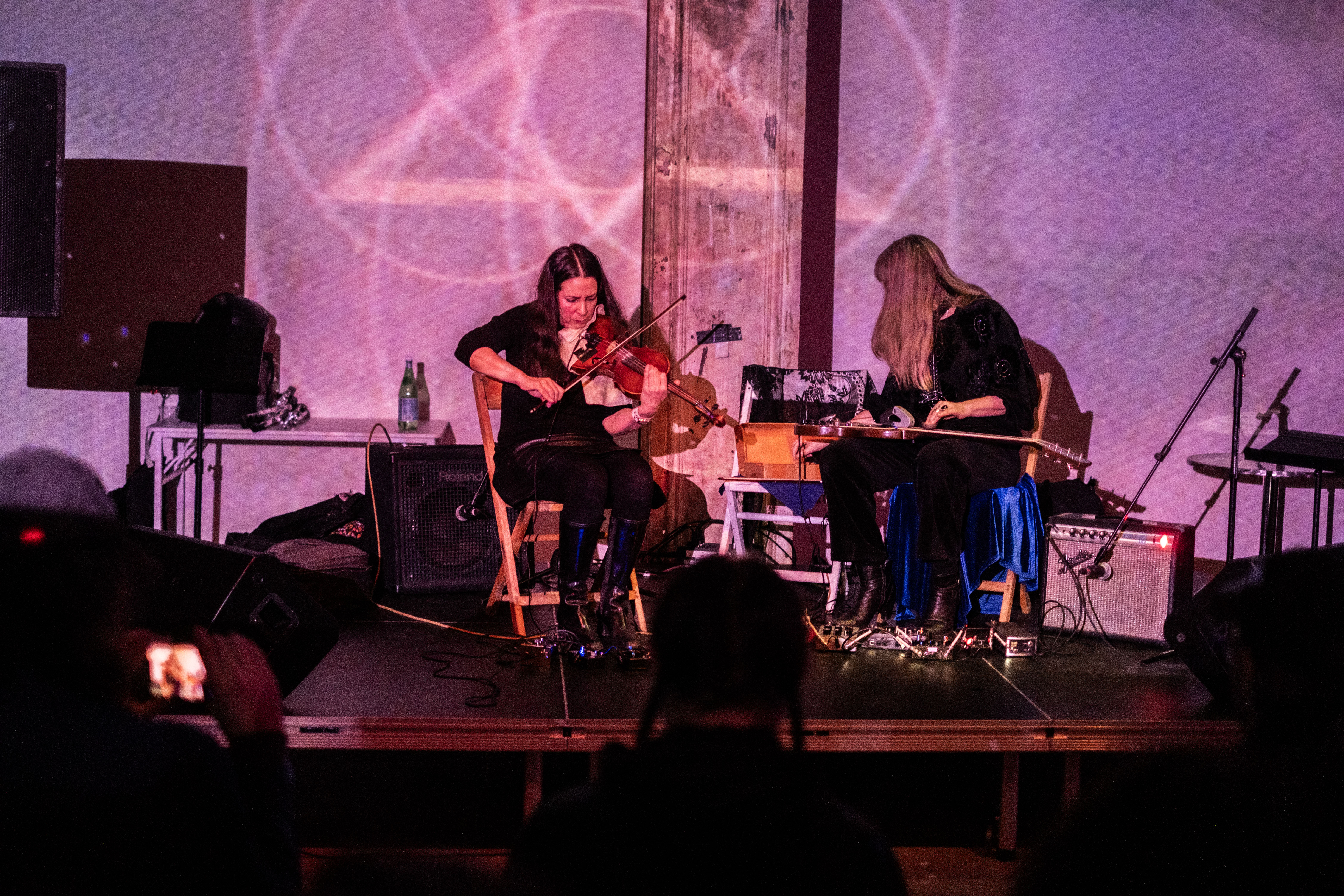 Color image of two performers on stage, both seated in chairs. The musician on the left plays a violin while the musician on the right plays a guitar that is placed across their lap. Each musician has several pedals placed on the floor that they control with their feet. Two amps flank the pair of musicians. The musicians and the entire stage is cast in a light pinkish purple light emitted from a projector. A design of overlapping circles with a triangle in the center are projected onto the back wall.