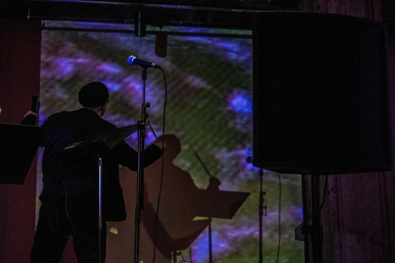 Color image of a performer standing in the midground of the image with their back towards the audience. They are dressed in all black including a black beret. Both of their arms are raised with a bend at the elbows. In their right hand, they hold a straight object, perhaps a conductor