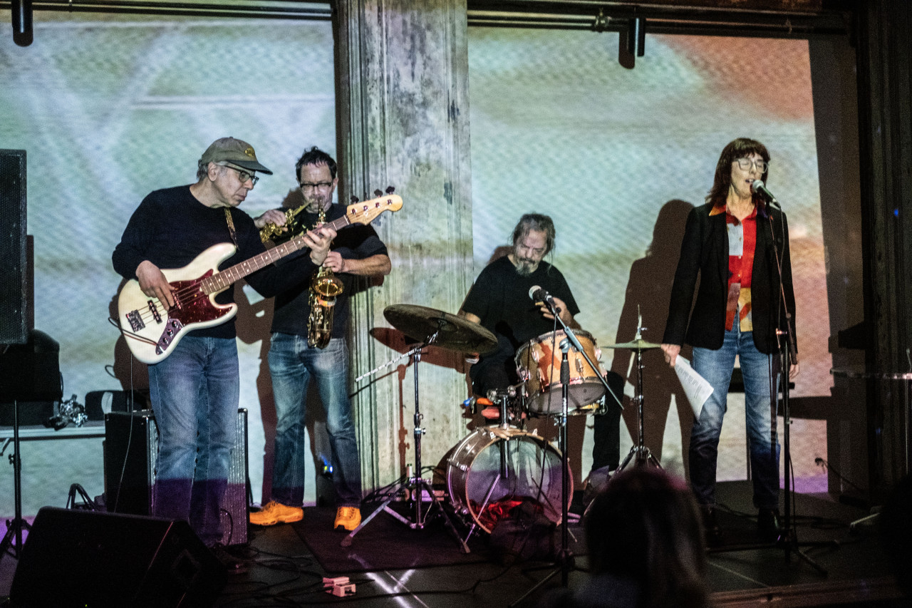 Color image of a four-person band on stage. The left-most musician wears a green baseball cap, black tee-shirt and jeans while playing a guitar. Slightly behind him to the right, a musician in a black tee shirt and blue jeans is seemingly playing an alto and tenor saxophone at the same time. To the right of the saxophonist, a musician with grey hair, mustache and beard plays the drums. To the right of the drummer, closer to the front of the stage, a singer stand upright, vocalizing into the microphone with their eyes closed. The singer grasps a sheet of paper in their right hand. The entire stage and back wall is illuminated by bright bluish-green light emitted from a projector. 