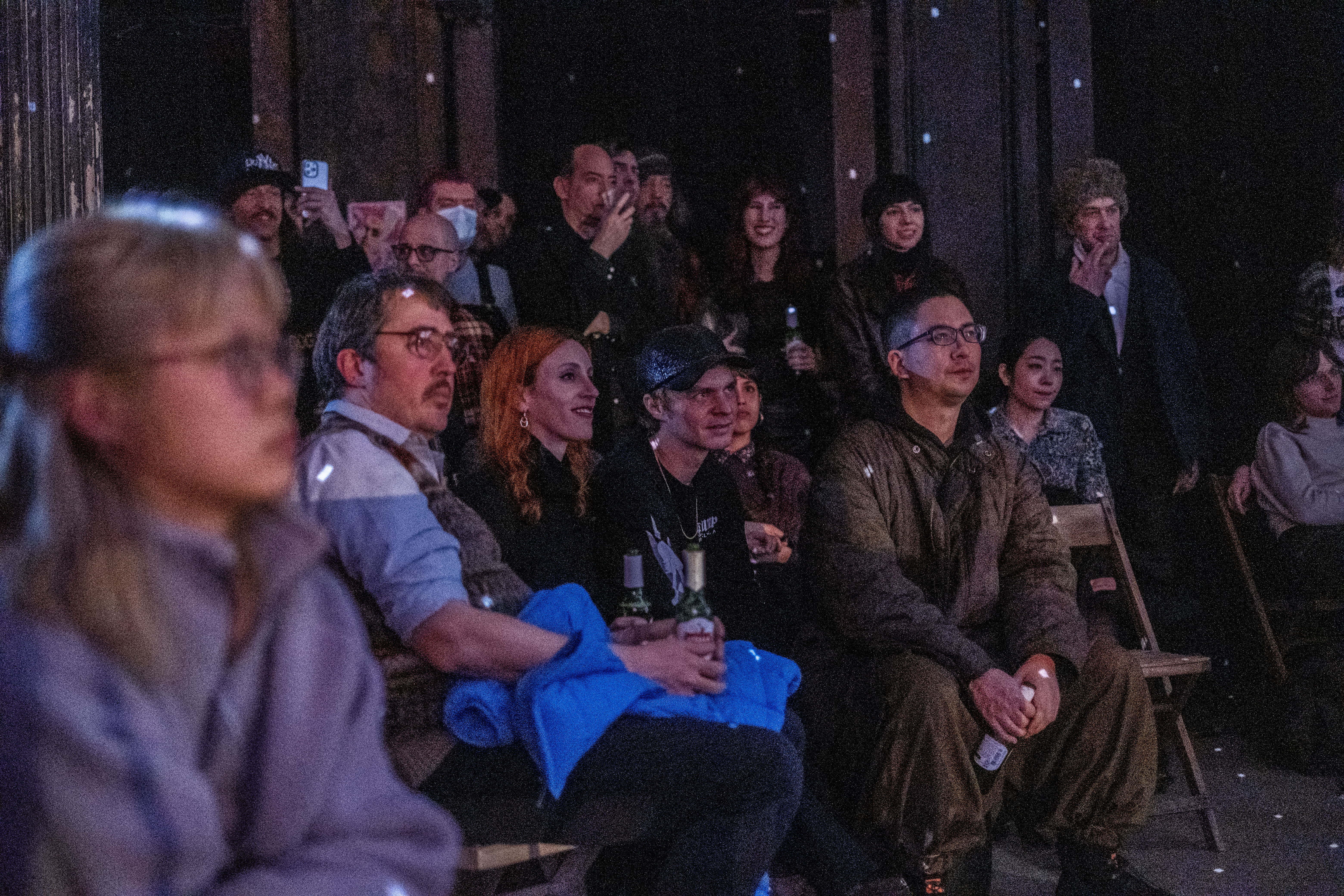Color image of the audience. In the foreground, audience members are seated in chairs and in the background, folks stand and watch the performance. Almost all of their eyes are attuned towards the stage, which is out of frame to the right. Many of the audience members wear smiling, joyful faces as they watch the show. The audience sits in the dark of the seating area, small specks of bright white light reflecting from a disco ball scatter on their bodies and the walls behind them. 