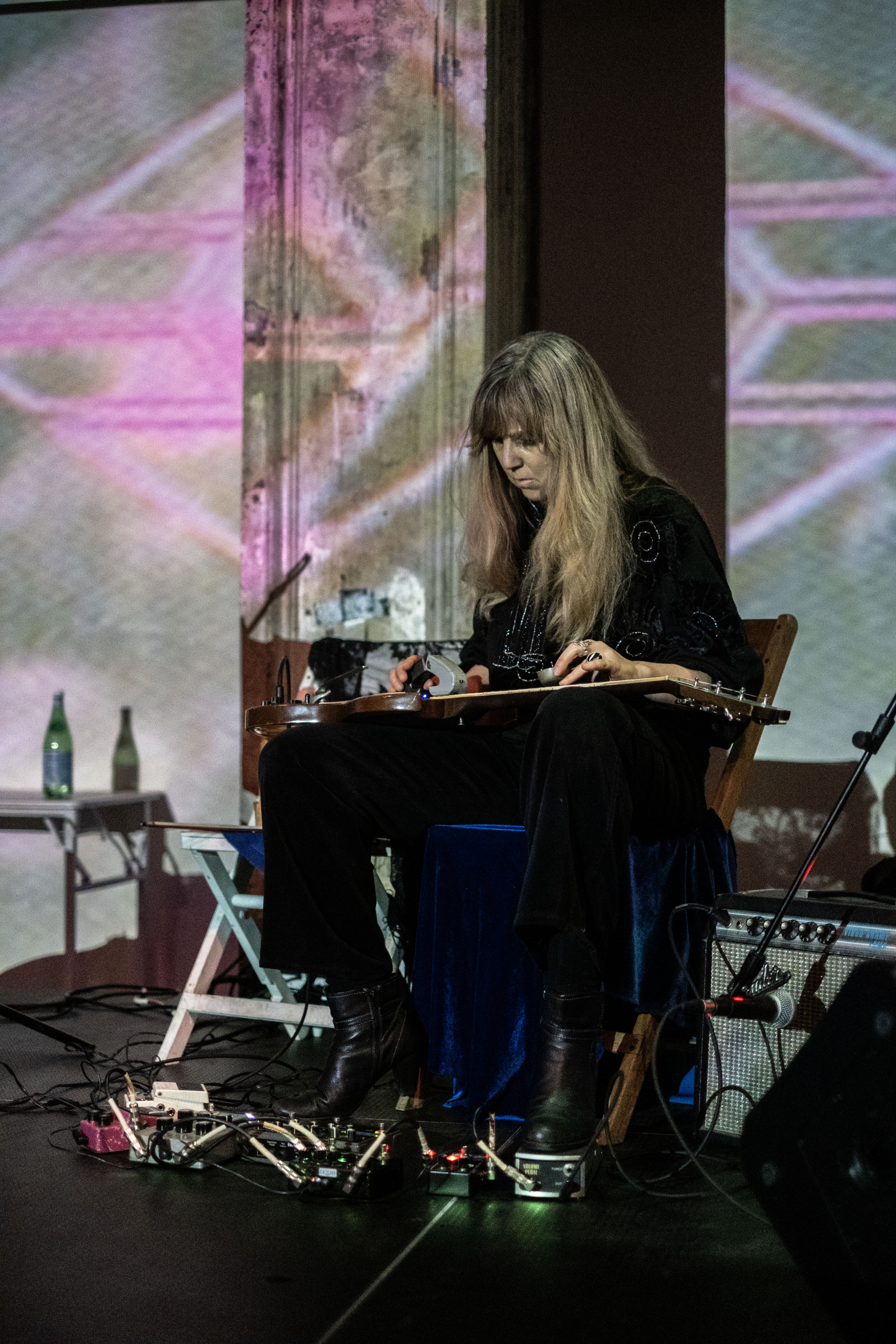 Color image of a musician on stage, seated in a chair, holding an instrument that appears to be a guitar across their lap. To their right, a microphone is positioned close to an amp. Their feet control several pedals placed on the floor. Their long blonde-brown hair falls down over their chest and back as they gaze down in deep focus at the instrument in their lap. Behind the musician, a diamond-shaped, geometric pattern, outlined in bright pink is projected on the back wall. 