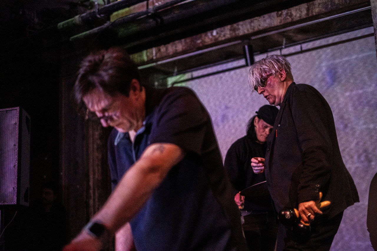 Color image of three performers on stage. In the left foreground, a blurred figure in a black polo leans over, their arms stretched out, presumably towards an instrument placed on a table. In the midground, a tall performer dressed in all black with shaggy silver head and sunglasses is caught mid-motion with a pair of macarenas in their left hand. Behind the silver-haired performer, we catch a glimpse of a drummer in a black hat, their face turned down towards the cymbal they are about to strike. Behind all three performers, the back wall is illuminated in a white light. 