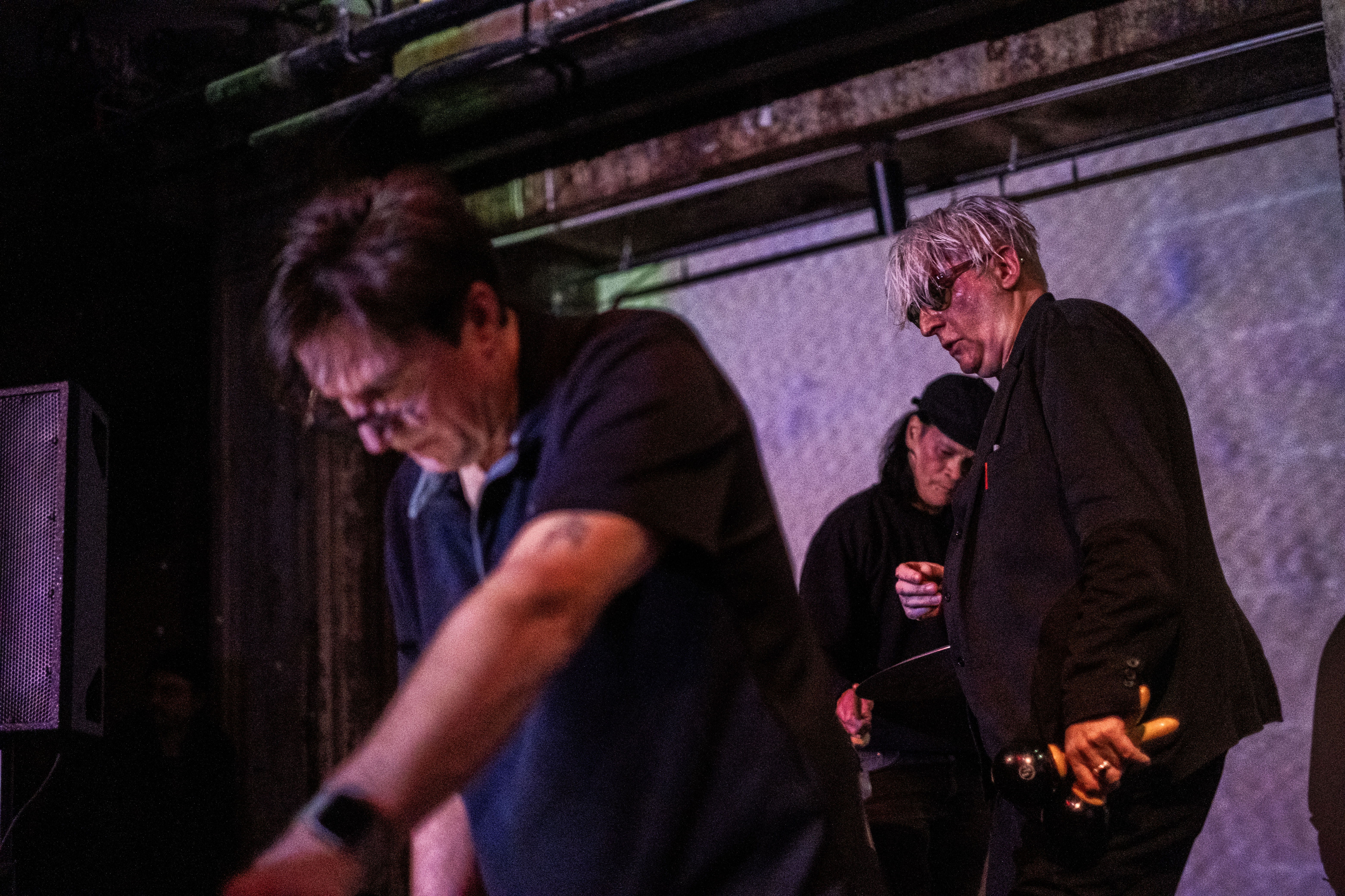 Color image of three performers on stage. In the left foreground, a blurred figure in a black polo leans over, their arms stretched out, presumably towards an instrument placed on a table. In the midground, a tall performer dressed in all black with shaggy silver head and sunglasses is caught mid-motion with a pair of macarenas in their left hand. Behind the silver-haired performer, we catch a glimpse of a drummer in a black hat, their face turned down towards the cymbal they are about to strike. Behind all three performers, the back wall is illuminated in a white light. 