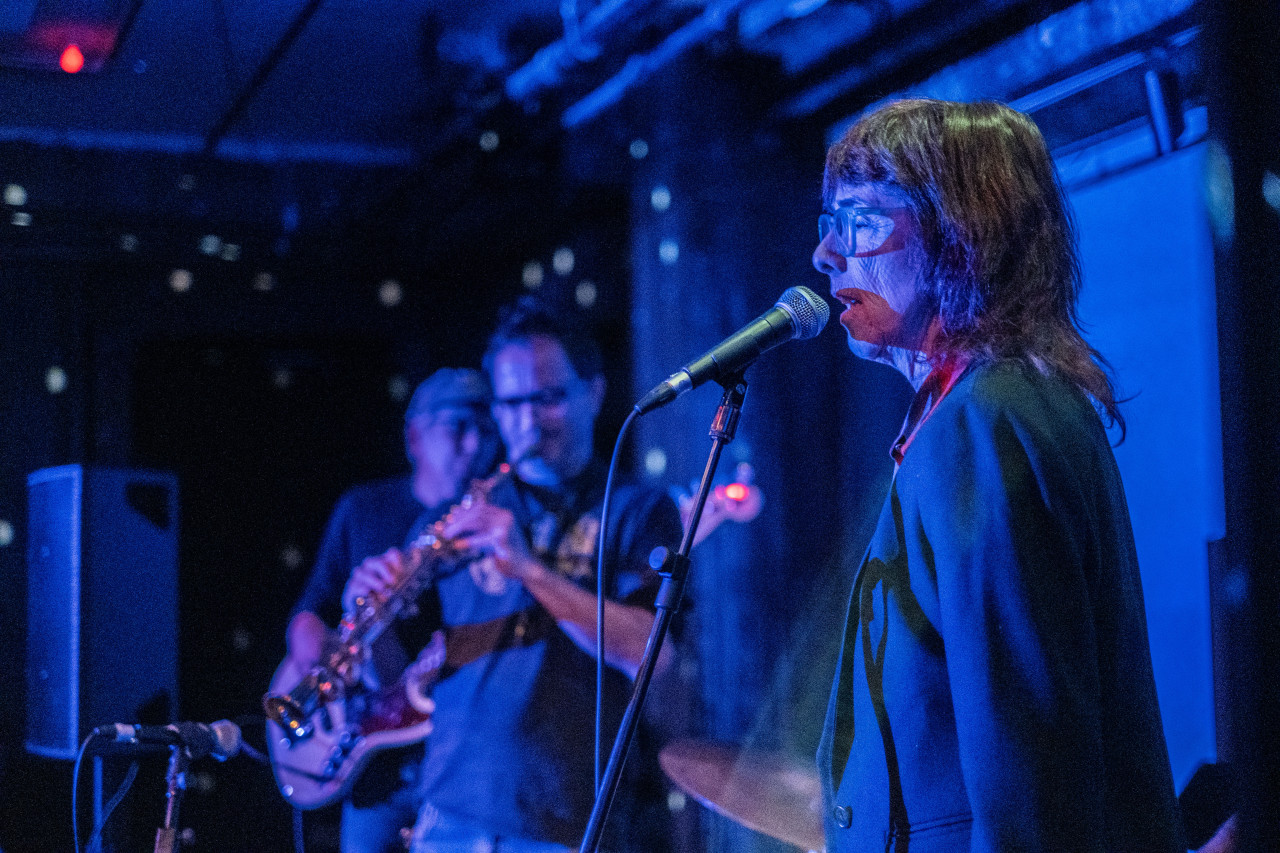 Color image of three performers on stage. In the right foreground, a performer with medium length dark hair and glasses vocalizes into a microphone. Blurred in the left background are two more performers. The one standing closest to the vocal artist plays a tenor saxophone instrument while the musician behind him plays a guitar. The entire trio and interior is cast in a cobalt blue light emitted from a projector. 
