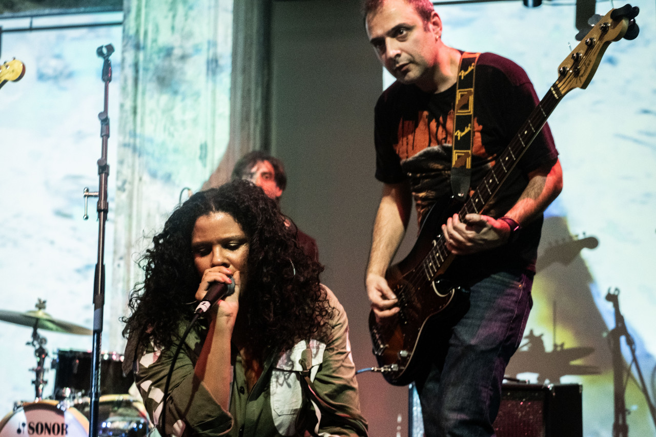 Close-up color photograph of a vocalist and a musician playing a bass at the edge of the stage. The vocalist has long curly black hair and wears an army-green cargo jacket. They are kneeling down as they hold the microphone up to the mouth. To the right of the vocalist, the bassist looks out to the crowd as picks the strings. The drums and the drummer are visible behind them. The wall behind the stage is illuminated by a bright white light emitted from a projector. 