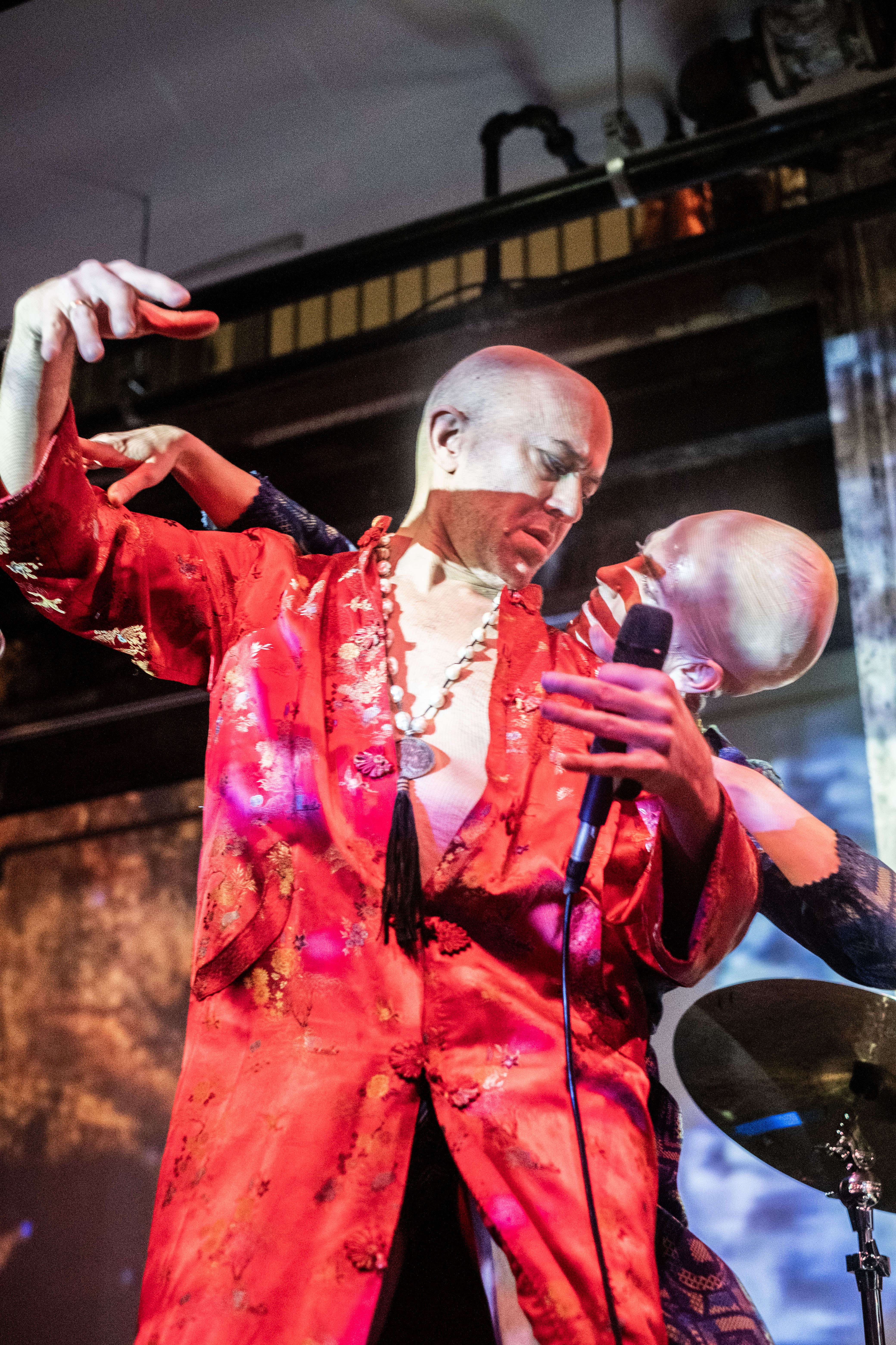 Close-up color image of two performers. Occupying most of the image is a performer in a red robe with only one button buttoned so the middle of their chest is exposed. A necklace with chunky white beads and a silver pendant with a black tassel hangs around their neck and down their bare chest. The performer's right hand is raised up, their head turned down, looking over their left shoulder to the second performer who stands behind them. The performer dressed in red with a bald head holds a microphone in their left hand. On the red-dressed performer's left shoulder, another performer, wearing a bald cap looks up at them. Their right hand is raised, touching the red performer's forearm. Their faces and the wall behind them is illuminated by the bright white light of the projected image.  --