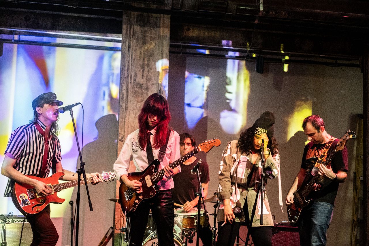 Color photograph of a five person band on stage. To the left, a musician playing a guitar wears a black and white vertically striped shirt and a black conductor hat. They sing into a microphone. To the left of the musician playing guitar, another musician holds a guitar while wearing a white button up and black leather pants. Behind the guitarist, a musician in a black tee shirt plays a set of drums. On the right side of the image, a musician plays a bass in a black tee shirt and jeans. To the left of the bassists, a vocalist in a cargo jacket and black beanie prepares to vocalize into the microphone. The stage and the wall behind the stage are illuminated by a colorful projection. Some of the band