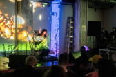 Color image of a seated musician playing a tenor saxophone on a stage. In the foreground of the image, there is the blurred back of the heads of seated audience members. In the center midground of the image, the musician is basked in green and yellow light being emitted from a projector. The projection covers the back wall behind the musician and the stage. 