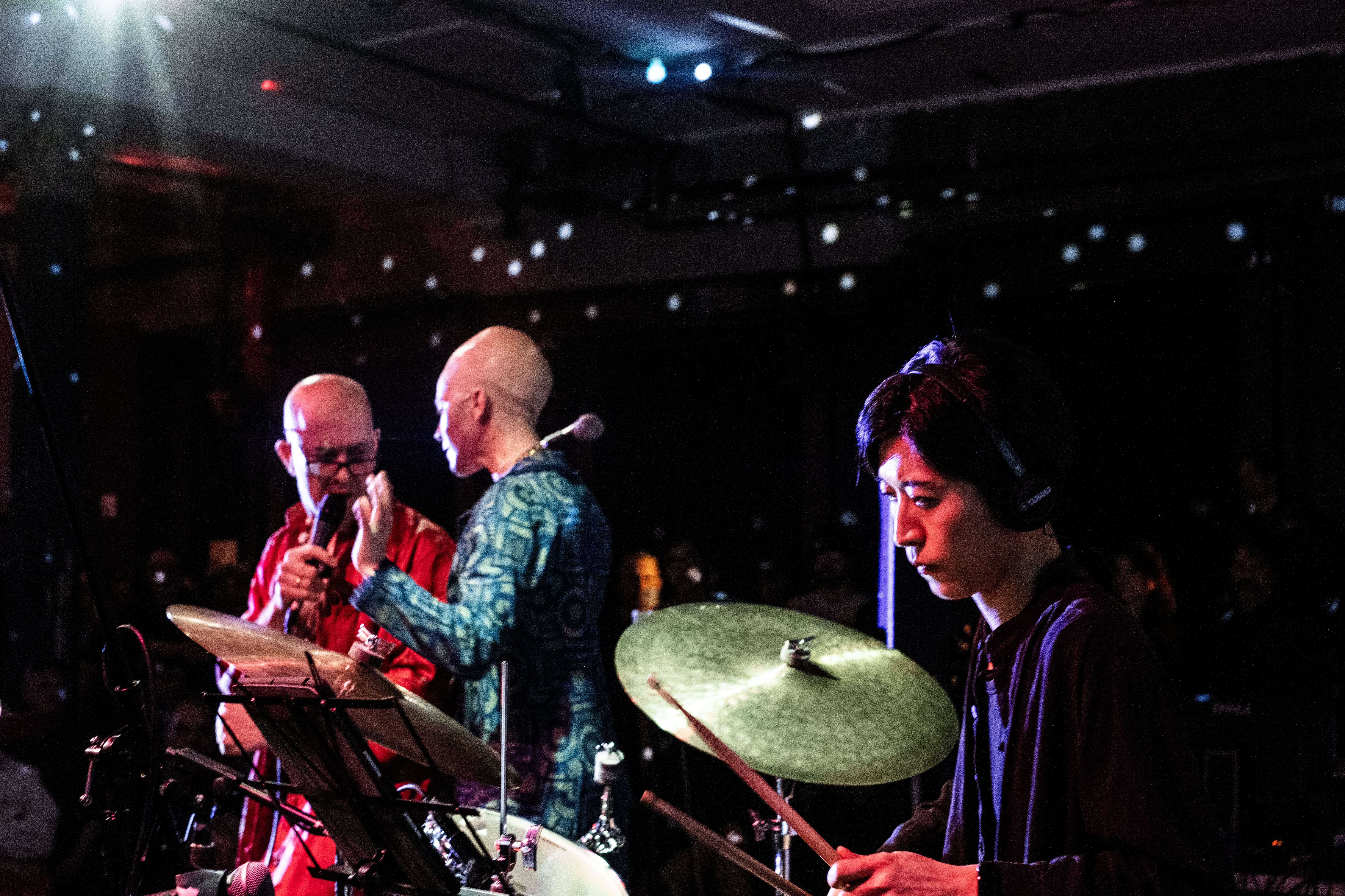 Color image of three performers on stage. In the foreground, in the lower right corner a musician wearing headphones plays drums. To the left hand side in the midground, two bald performers look at one another. One of the performers wears a red robe and holds a microphone. The other performer wears a blue tunic and faces the performer in the red robe. Stage lights shine down on all three performers. 