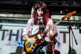 Close-up color photograph of a musician holding a guitar. The musician wears a white-button up with a black tie and black leather pants. Medium-length red-and-black streaked hair falls around their face as they look down at the guitar. The musician and the wall behind them is illuminated by a projected image that is mostly white, but features large black letters at the top of the projection. 