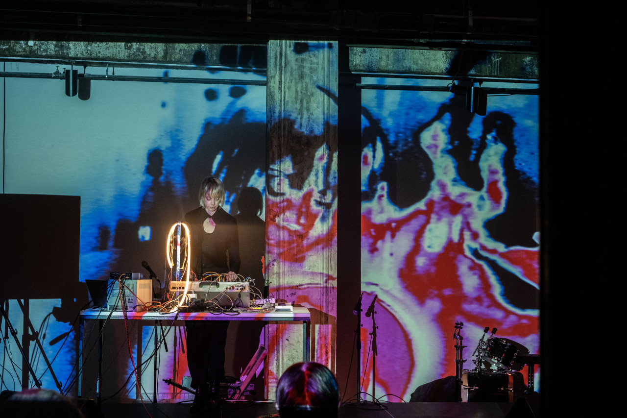 Color image of a musician standing behind a long table on a stage. The musician plays a large soundboard placed on the table. The musician wears a black long sleeve top and looks down at the soundboard as they play. They have short blond hair with bangs. A colorful image, almost like a close-up shot of a cell rendered in pink, red, blue and black is projected onto the stage and the wall behind the stage. A shadow of the musician appears on the back wall. 