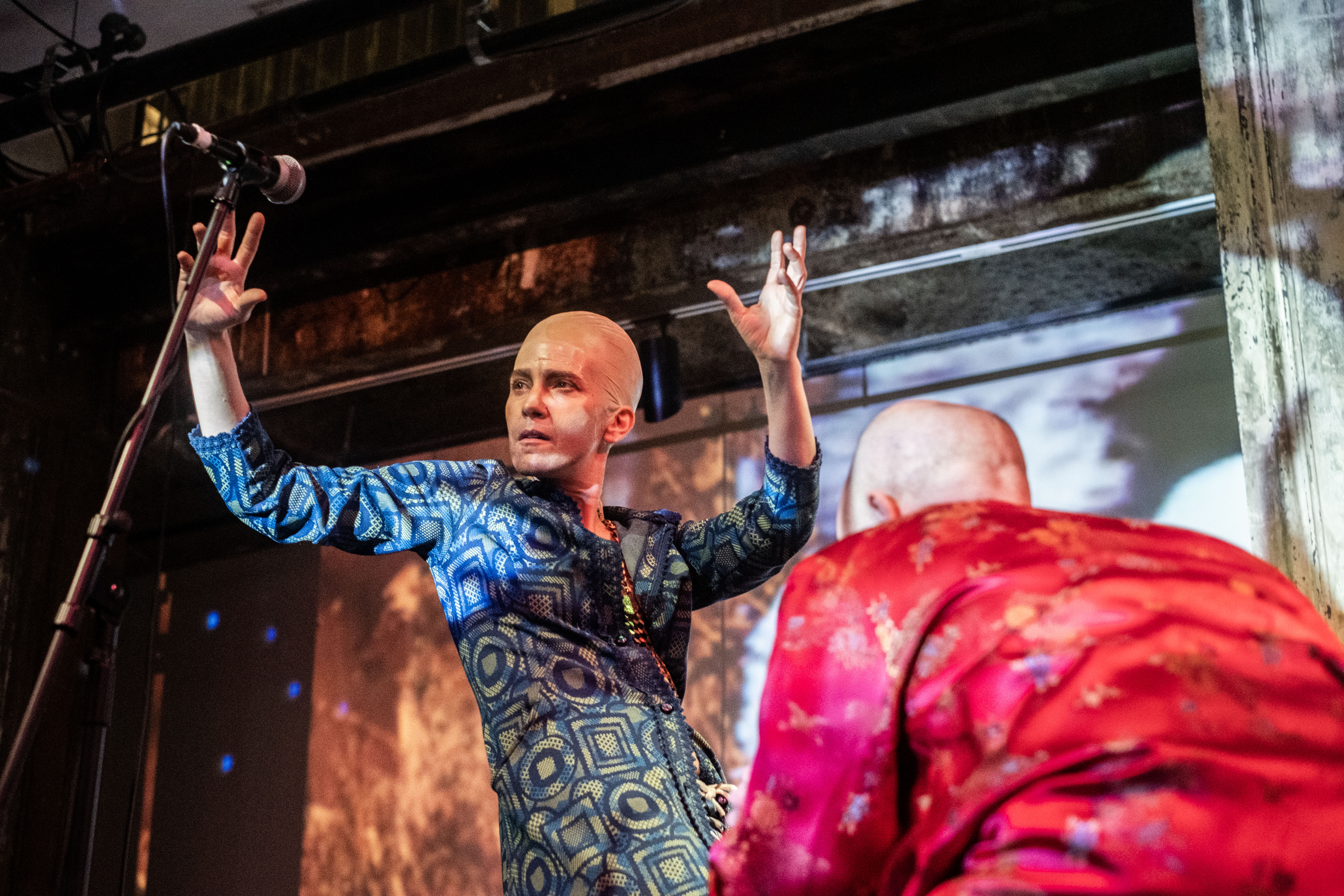 Color image a performer in a bald cap with both of their hands raised with elbows at shoulder height. The performer occupies the center of the image wearing a blue dress covered in a square and circle pattern. From the left, bottom corner, a microphone on a stand rises to meet the performer in the center. In the lower, right hand corner, another bald performer, dressed in a red top bends down towards the performer in the center of the image. The wall behind both the performers is filled with a projected image in hues of yellow, brown and white. 