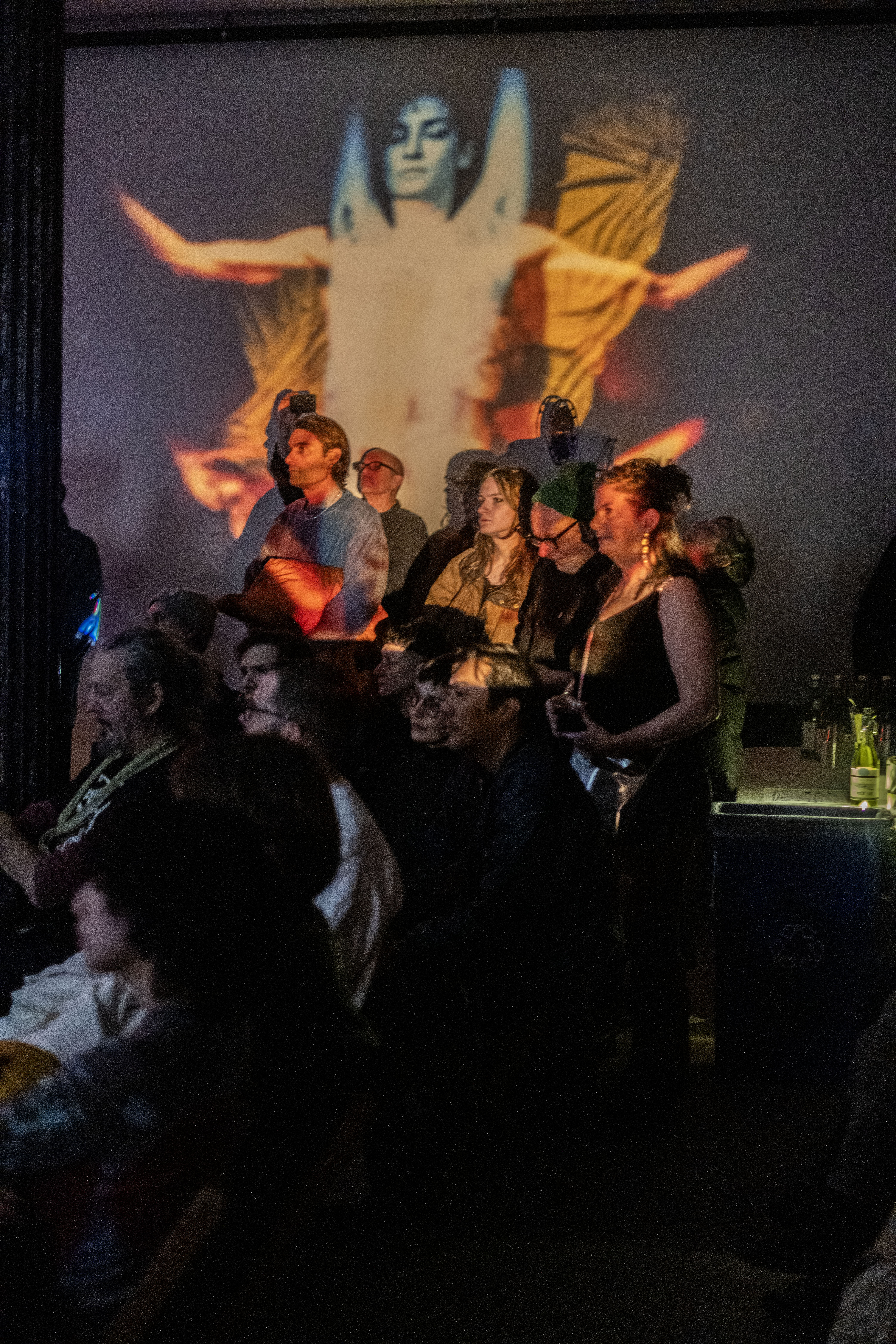 Color image of audience members watching a performance out of the left side of the frame. On the left side of the image, there are two rows of seated audience members in shadow. In the center of the image, audience members stand against a wall. The upper half of the standing audience members is cast in orange and red light emitted from a projector. Behind the standing audience, an image of a figure rendered in red, yellow and blue is projected on the wall. On the right edge of the image, there is a table with clear glasses and bottles of wine. 