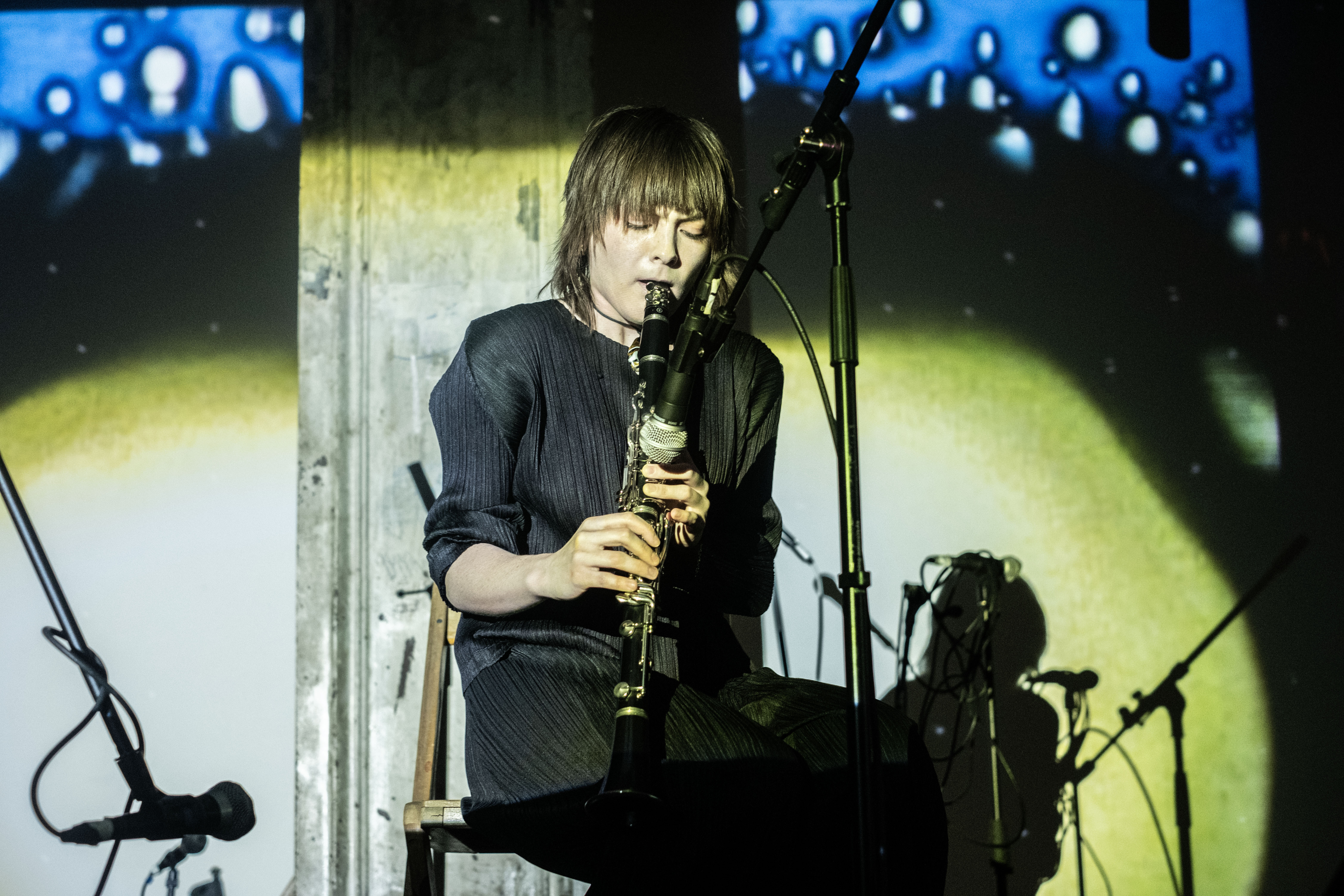 Color image of a seated musician playing an oboe into a microphone. The musician is dressed in all black and has medium length black hair. The image catches the musician with their eyes downturned, just a moment before blowing air into the oboe. The musician and the back wall are flooded with a circle of yellow light, in the top corners, the yellow turns into blue. 