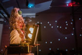 Portrait view of solo performer, next to a small suitcase opening to show light bulbs and several small wires. The wall behind performer shows the lights from and the shadow of a disco ball. The performer has thick red lipstick and an eyeball on top of their head amidst long blonde hair.