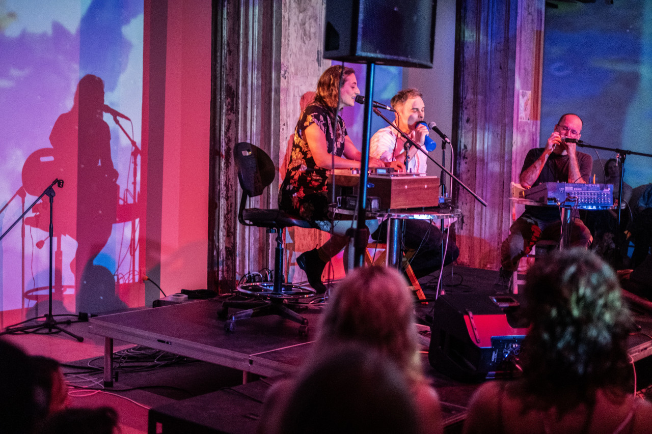 View of 3 performers on stage from center left. Performers pictured in front of soft pink, blue, and white light against a wall. Leftmost performers is seated, speaking into a microphone and manipulating a tape machine with hands. Performer in center is drinking from a blue water bottle. Rightmost performer is making sound through cupped hands into microphone. Sharply defined shadows against back wall.