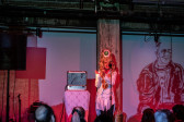 Solo performer on stage next to a small table with a silver metal box on top. The walls are bathed in soft pink and purple light, to the right of the performer an image of a man in the style of a lithograph is projected. The performer has a green eyeball attached to their head amidst long flowing blonde hair. 