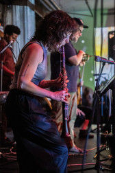 In the foreground a musician is playing a clarinet, slightly bent forward. Behind them are two other performers on stage at rest. The wall behind is lit with soft green light and the lights from a disco ball.