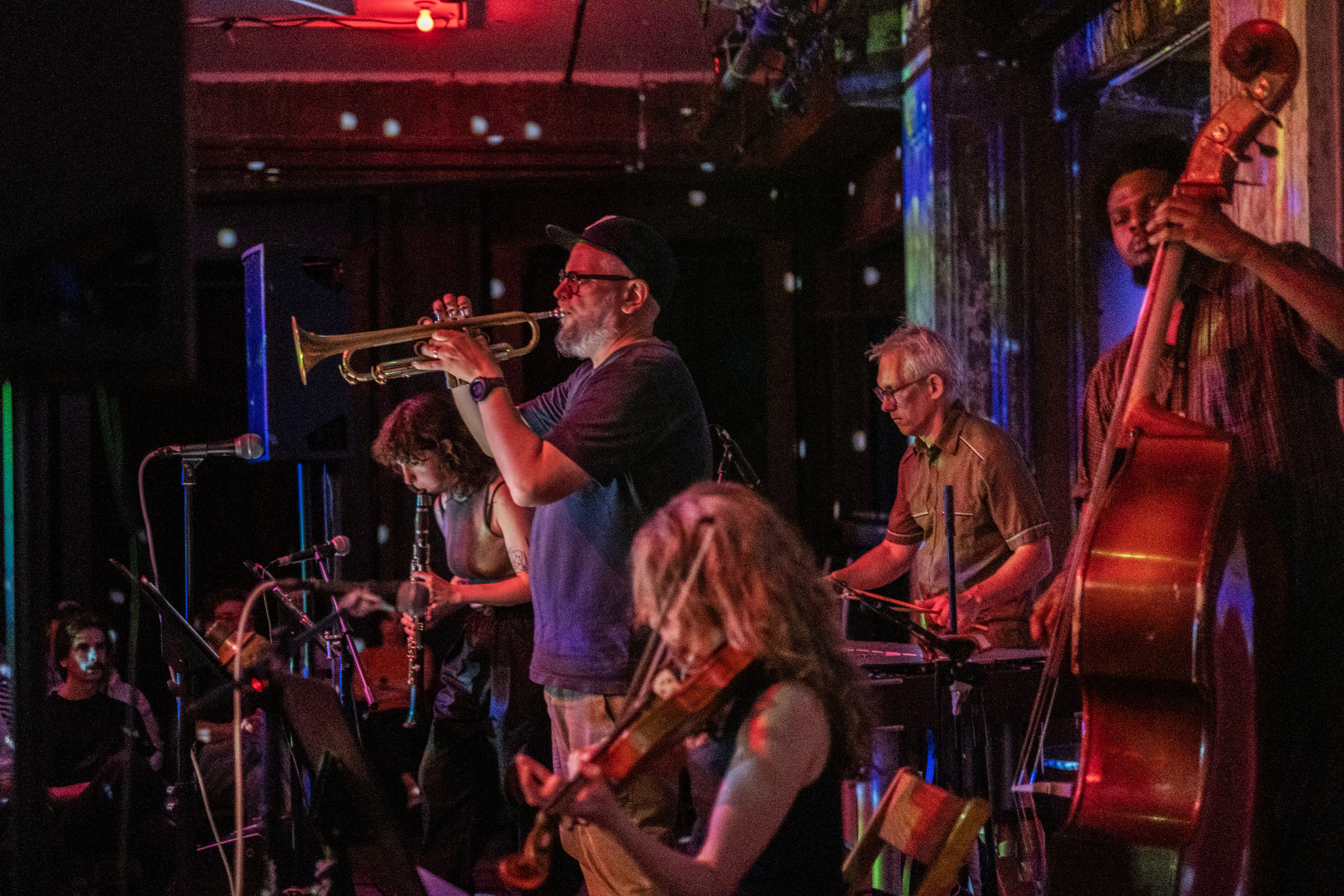 View from right of 5 musicians, with lights from a disco ball visible along the walls behind them. From left, musician playing a clarinet, behind which is a musician playing a vibraphone. In the center a musician playing a trumpet into a microphone. In the foreground are pictured musicians playing a cello and a violin. 