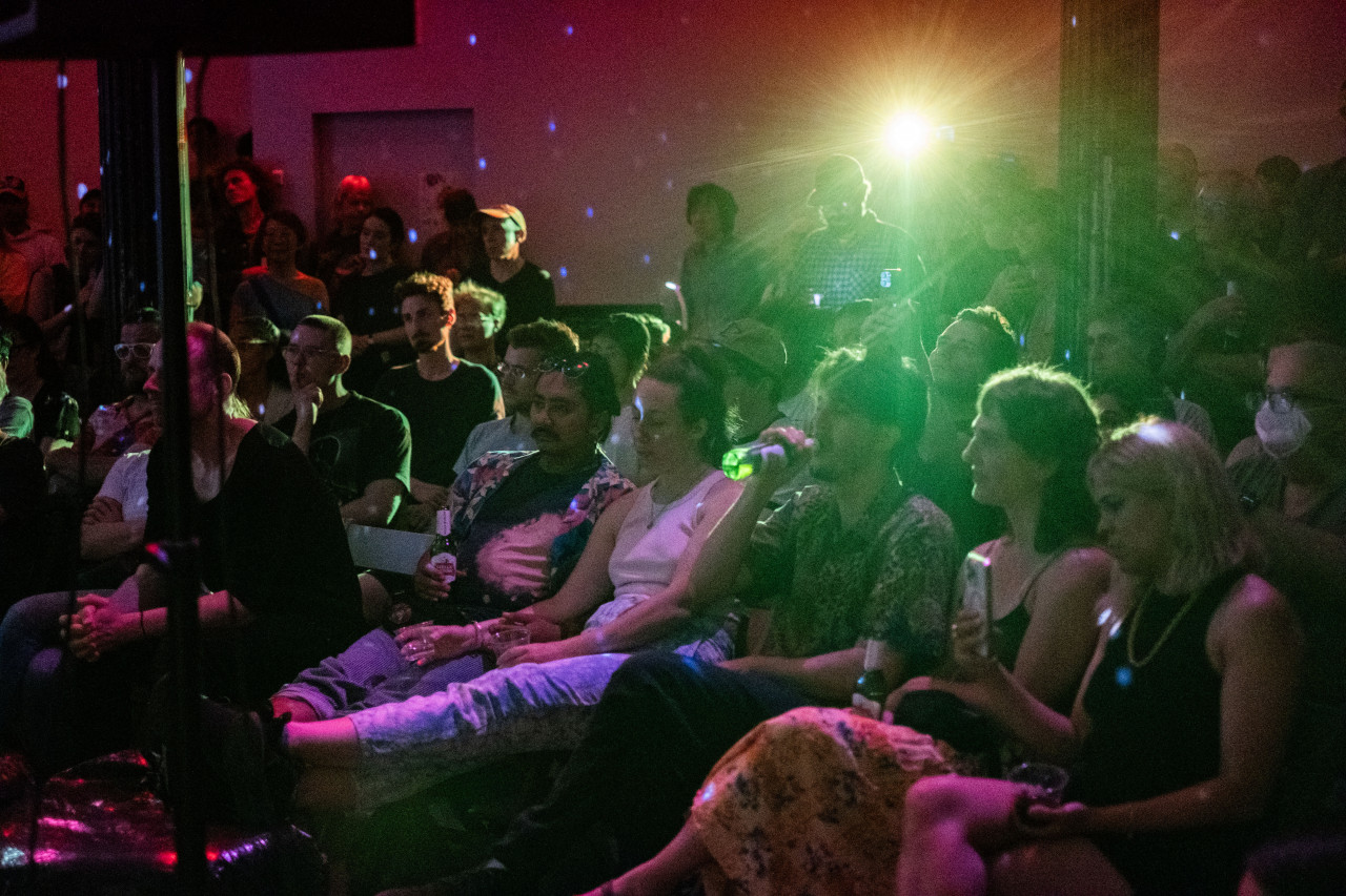 Audience members in a crowded room, seated, with a bright green light flaring from the center of the room. Around the back walls the lights from a disco ball can be seen.