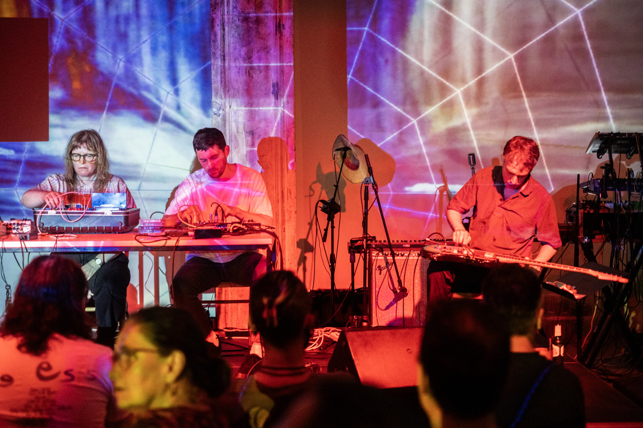 Three musicians sit on stage and play various electronic instruments. They are bathed in red, blue, pink, and purple light projections and a large scale image of a web. In the foreground audience members are lit with red and yellow lights.