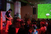 A solo musician standing on stage faces the audience and plays a small wooden instrument. They are bathed in red and pink light. They are looking down and wearing a headlamp. On the stage there are also multiple electronic instruments, cords, and a plant. A green image is projected on a side wall above the audience. The wall in the background is scattered with white dots reflected from a disco ball.