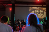 The audience faces the stage in the background, next to a projected graphic of an psychedelic-style eye and a red exit sign. The backs of two musicians are blurred in the foreground. The musicians are lit with soft blue and green lights. The room is dimly lit, and audience members are also tinted with multicolored lights.