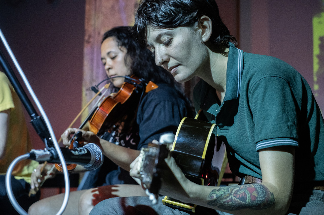 Close up of two musicians sitting side by side and looking down. The musician on the right plays the guitar and the musician on the left plays the violin. The wall in the background is purple tinted.