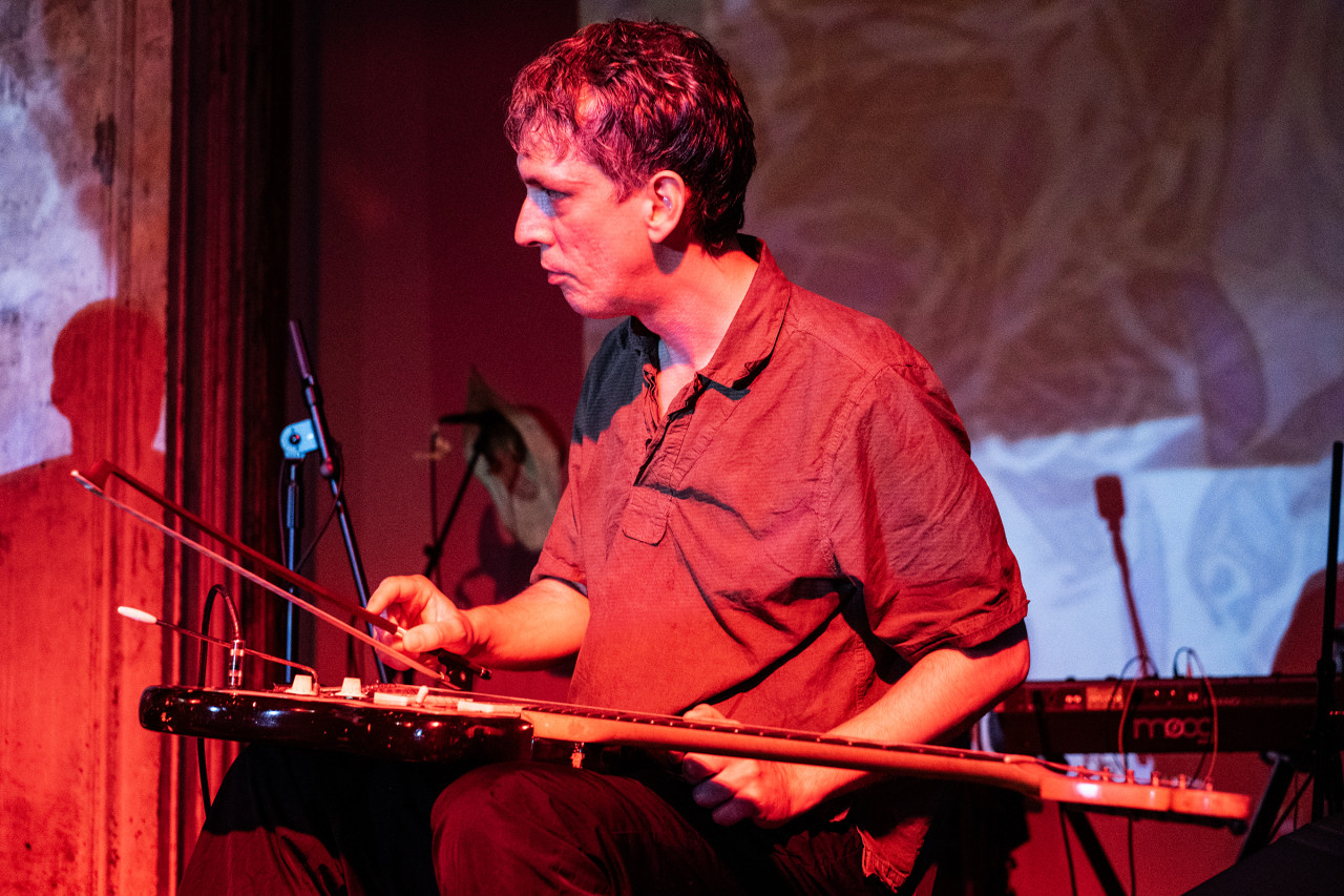 Close up of a solo musician sitting on stage playing a string instrument in their lap with a bow. They are looking to the side and they are covered in red light. The wall behind them is blue-tinted with patterned image projections.