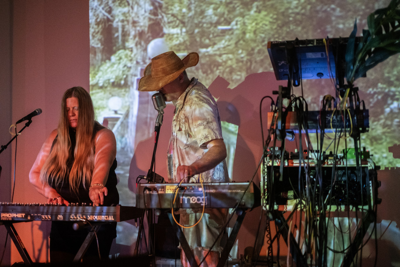 Two musicians standing on stage use electronic instruments. In the foreground there various more cords and audio instruments on stands. Both musicians have microphones and one is wearing a woven hat. The wall behind them is projected with images of trees, and a red light illuminates the left side of the frame.