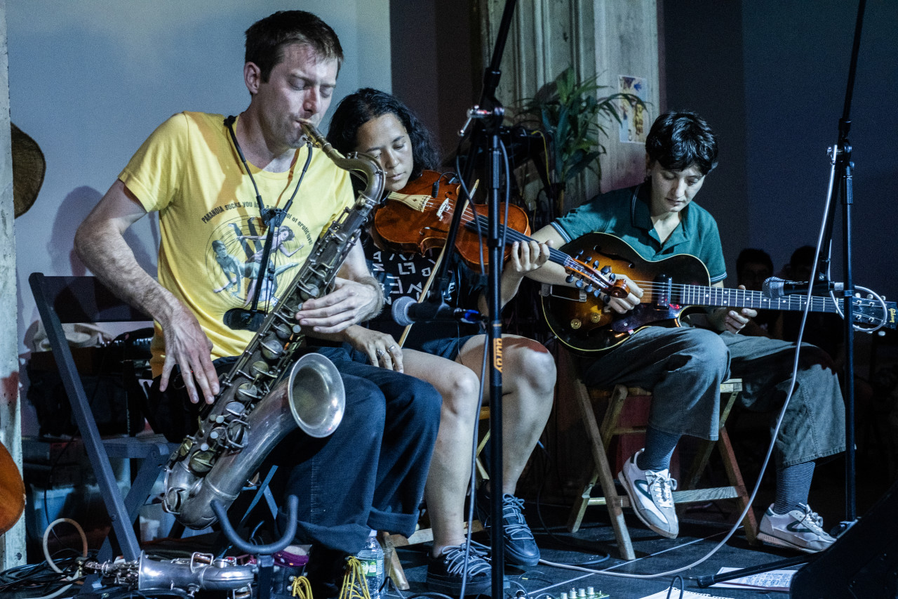 Three musicians sit on stage together. From left to right the instruments they are playing include saxophone, violin, and guitar. There are cords around the stage and a plant behind the performers.