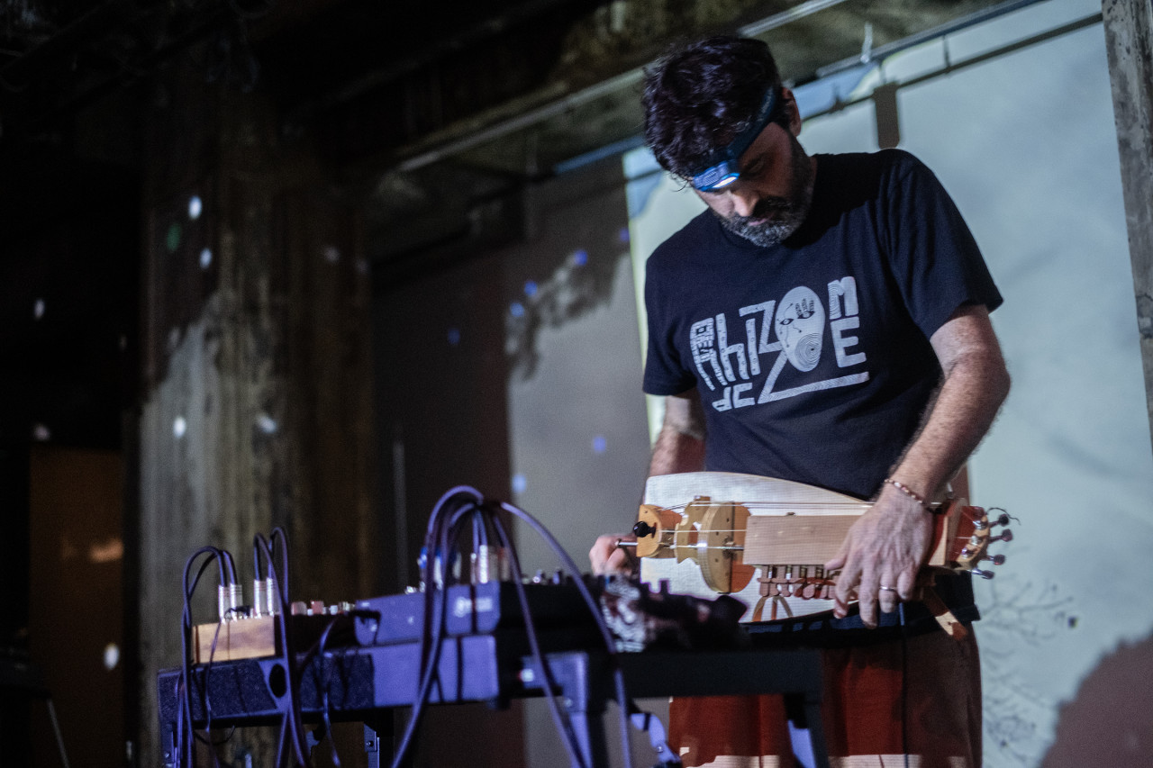 A musician standing on stage playing a small wooden instrument. They are looking down and wearing a headlamp. In front of them are multiple electronic instruments and cords. The wall in the background is scattered with white dots reflected from a disco ball.