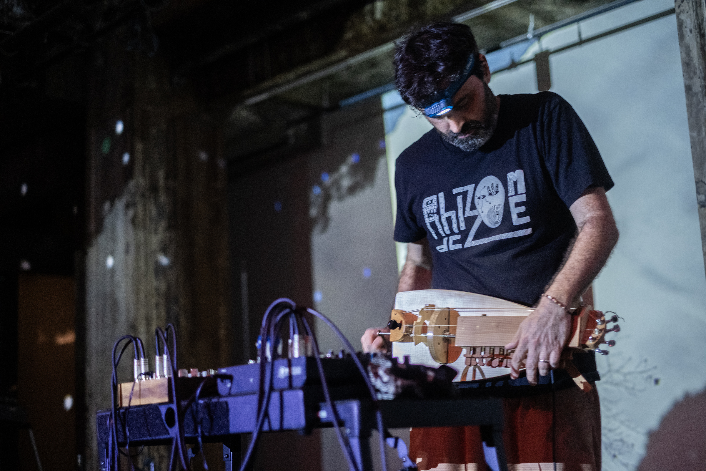 A musician standing on stage playing a small wooden instrument. They are looking down and wearing a headlamp. In front of them are multiple electronic instruments and cords. The wall in the background is scattered with white dots reflected from a disco ball.