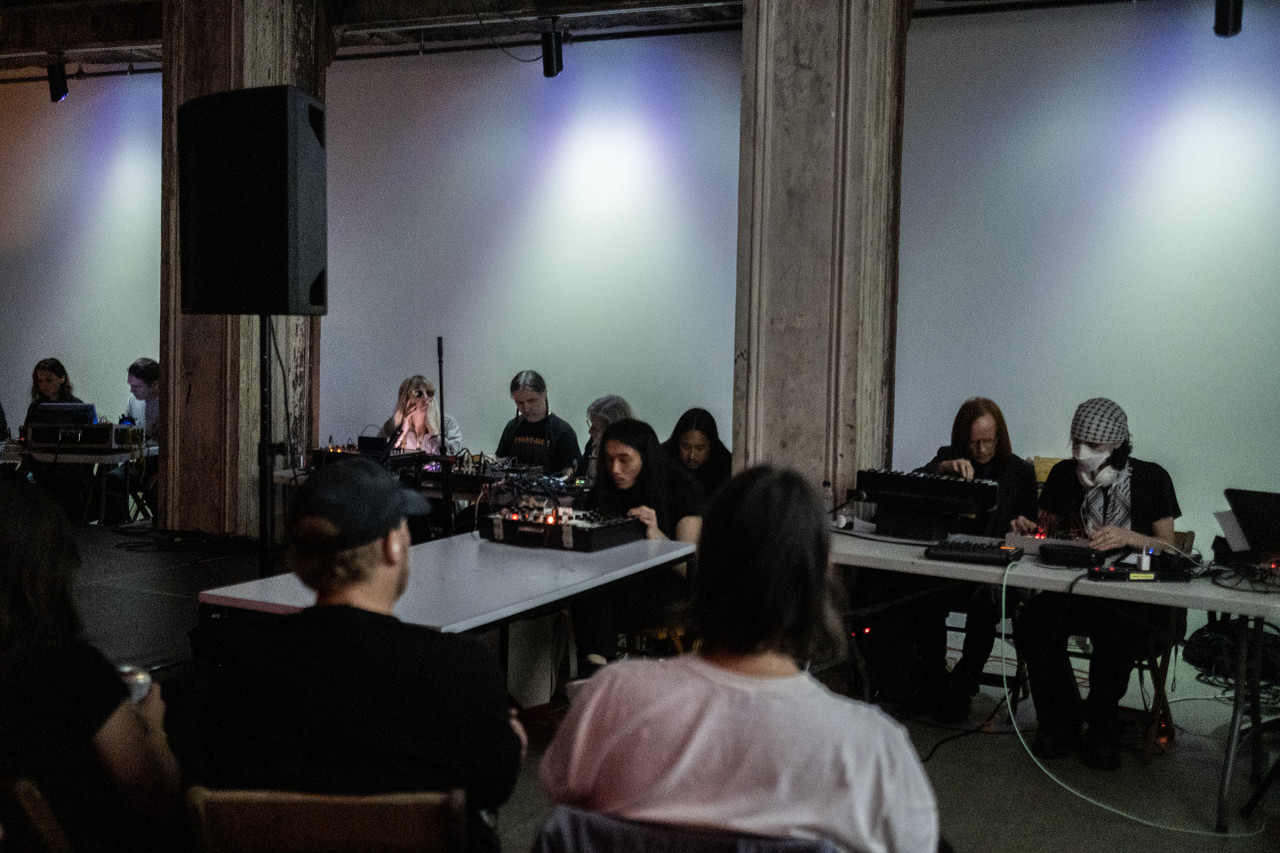 Nine musicians sitting side by side at tables use electronic instruments. The wall behind them is covered with blue tinted lights. There are a few audience members visible in the foreground.